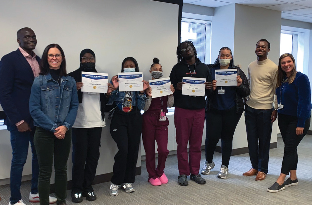 A group of nine people stands in a conference room; the five people in the middle hold up Certificates of Achievement for completing the Pathways Emerging Careers program.